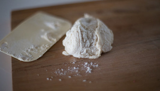 Flat bench scraper beside a ball of dough and a scatter of flour on a wooden counter.