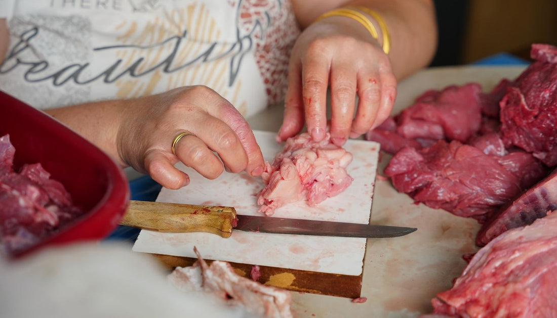 Hands trimming raw meat with a narrow knife on a white board, with more cuts of meat alongside.