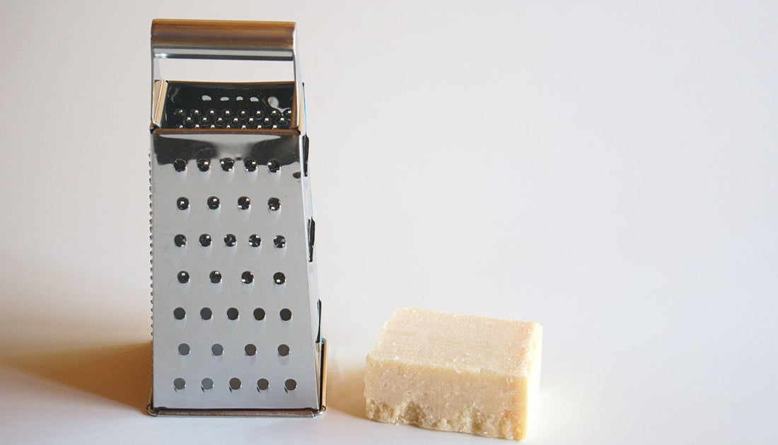 A stainless steel box grater standing next to a block of hard cheese on a white surface.