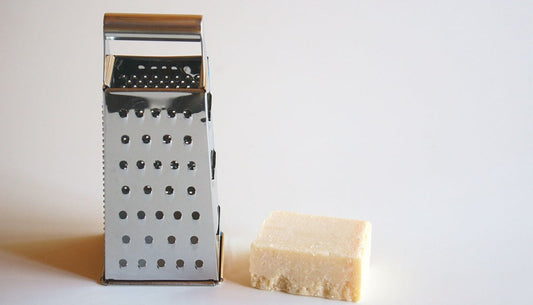 A stainless steel box grater standing next to a block of hard cheese on a white surface.