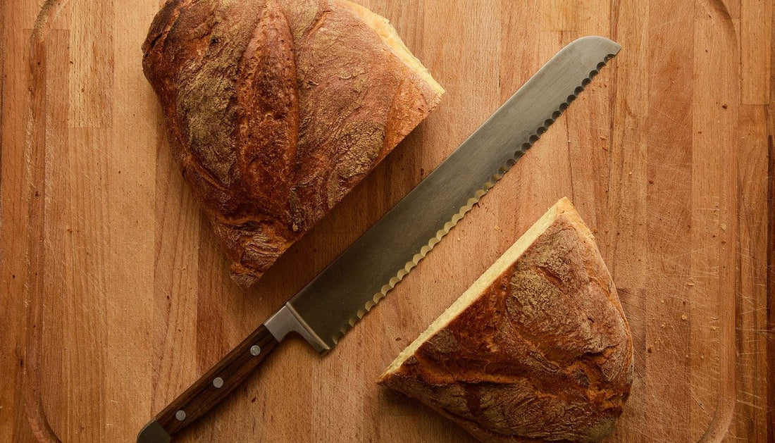 A serrated bread knife lying between two halves of a crusty loaf on a wooden cutting board.