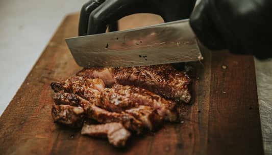 A gloved hand slicing seared steak with a cleaver on a wooden cutting board.