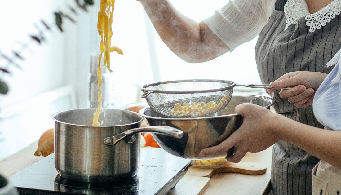 Two cooks pouring pasta from a pot into a metal colander held over a bowl.