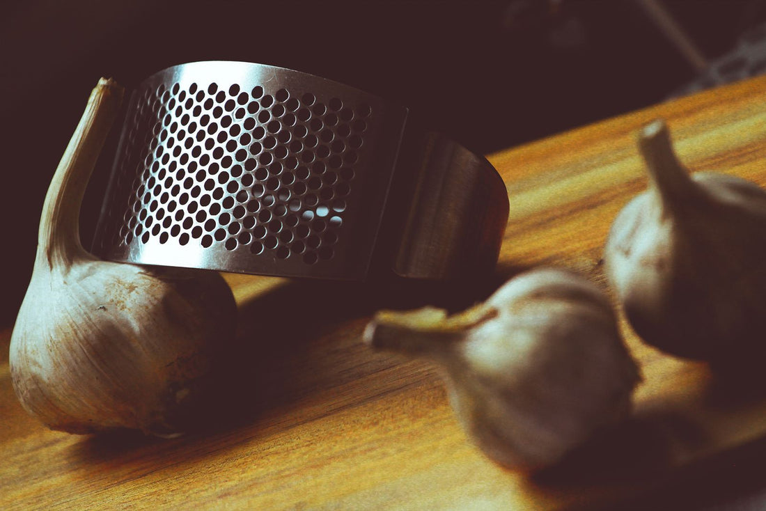 A stainless steel garlic press next to whole garlic bulbs on a wooden board.