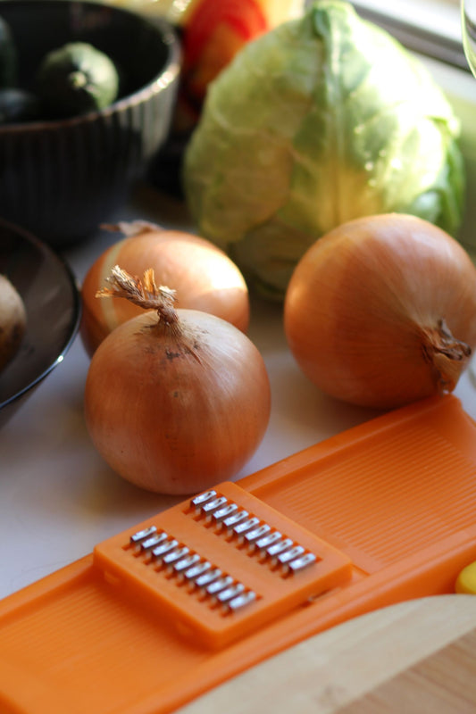An orange vegetable slicer next to onions and a cabbage on a kitchen counter.
