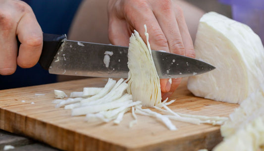 A cook slicing a head of white cabbage into fine shreds with a large knife on a wooden board.