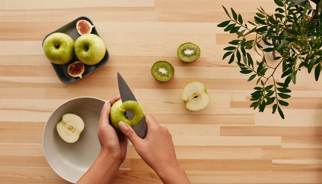Hands peeling a green apple with a small paring knife over a bowl on a wooden counter.