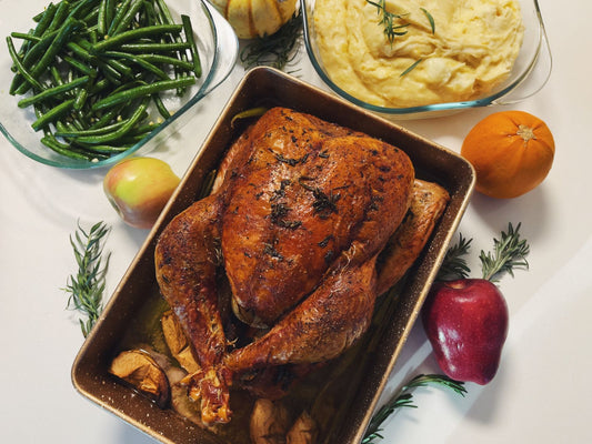 A roast turkey in a dark roasting pan surrounded by green beans, mashed potatoes, and fruit.