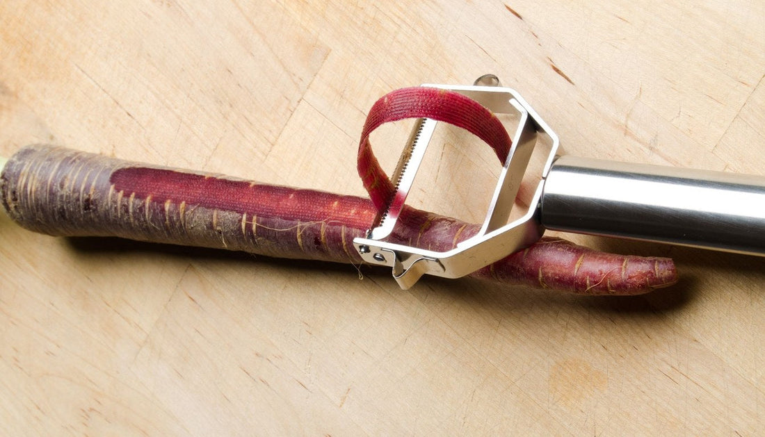 Y peeler taking a strip of skin off a purple carrot on a wooden board.
