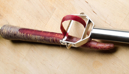 Y peeler taking a strip of skin off a purple carrot on a wooden board.