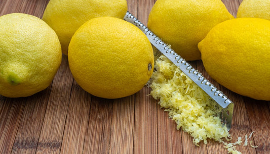 A long rasp-style grater beside whole lemons and a pile of fresh lemon zest on a wooden table.