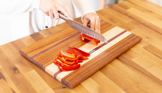 Hands slicing red bell pepper with a santoku on a striped edge grain hardwood cutting board.