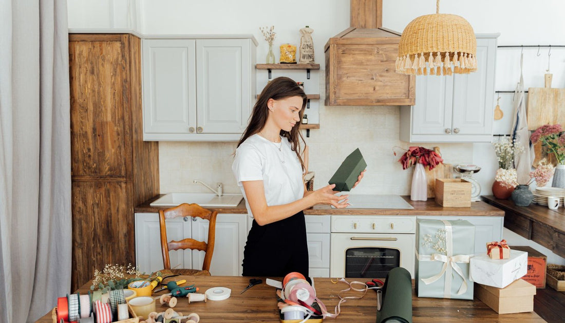 Woman wrapping gifts on a kitchen table with ribbons and boxes in a bright kitchen.