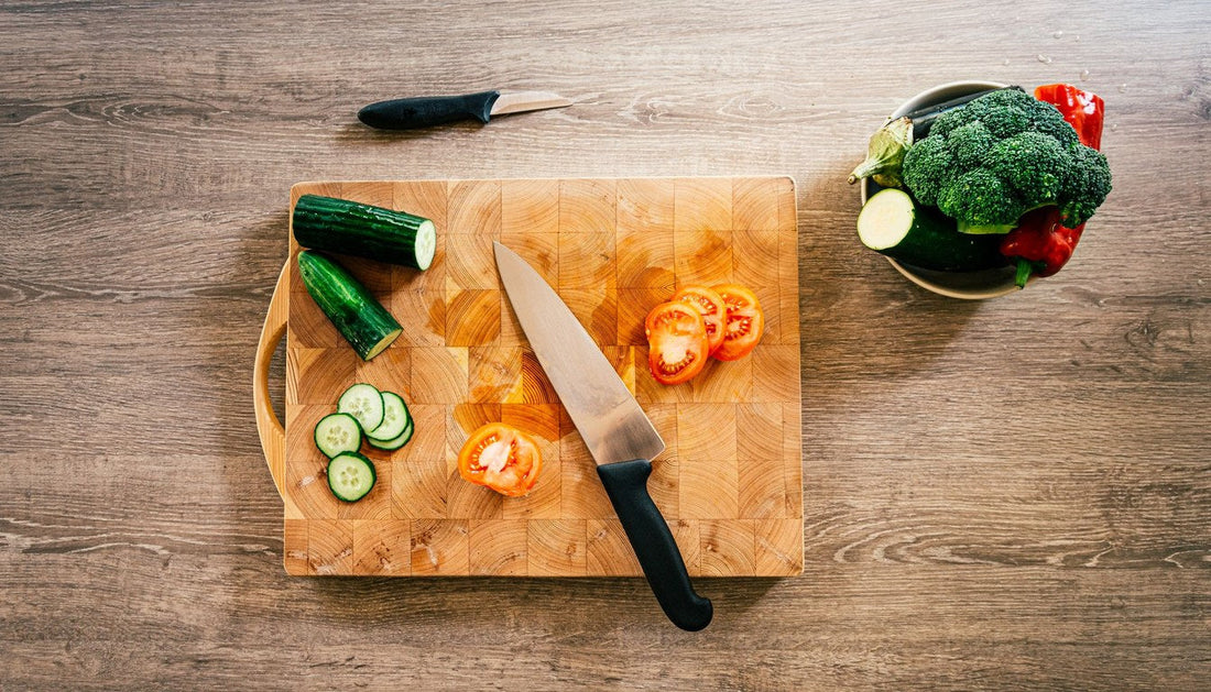 An end grain wooden cutting board with sliced cucumber, tomato, and a chef's knife on a wooden table.