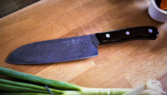 Santoku knife with a patterned steel blade resting on a wooden cutting board beside green onions.