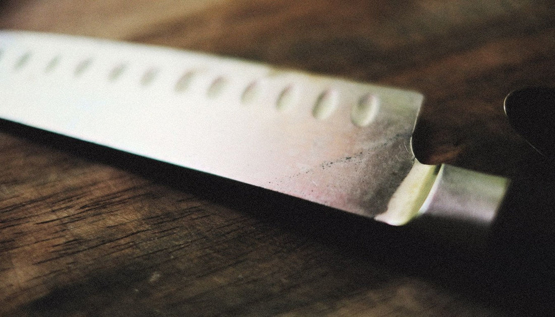 A chef's knife with a hollow-ground blade resting on a dark wooden surface.