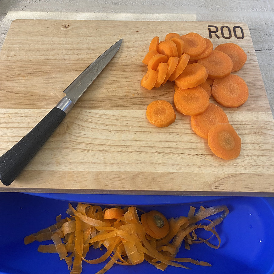 Sliced carrots on a cutting board, with the carrot peels already caught in the blue ROO mat's pouch below it